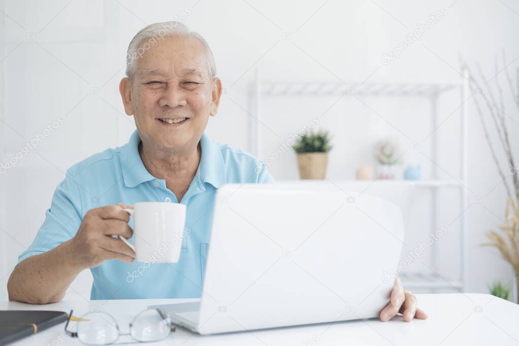 Happy senior man working on laptop and holding coffee cup at his home