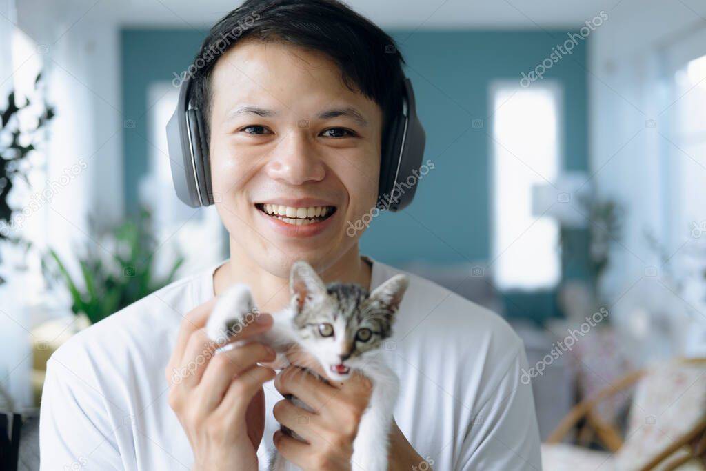 Head shot portrait attractive man holding cute kitten, making video call, using computer, sitting in living room, happy young male smiling, look at camera.