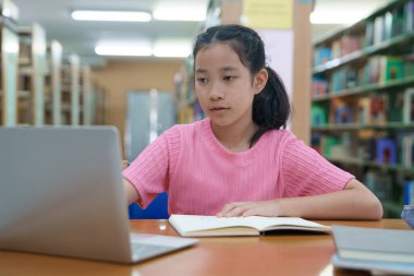 Çevrimiçi eğitim, e-öğrenme. Mutlu Asyalı ilkokul kızı şık günlük kıyafetler içinde, uzaktan çalışıyor, dizüstü bilgisayar kullanıyor, online dersler dinliyor, kütüphanede otururken notlar alıyor..