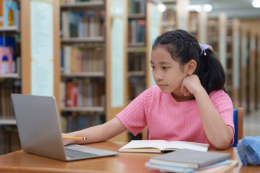 Çevrimiçi eğitim, e-öğrenme. Mutlu Asyalı ilkokul kızı şık günlük kıyafetler içinde, uzaktan çalışıyor, dizüstü bilgisayar kullanıyor, online dersler dinliyor, kütüphanede otururken notlar alıyor..