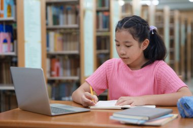 Çevrimiçi eğitim, e-öğrenme. Mutlu Asyalı ilkokul kızı şık günlük kıyafetler içinde, uzaktan çalışıyor, dizüstü bilgisayar kullanıyor, online dersler dinliyor, kütüphanede otururken notlar alıyor..