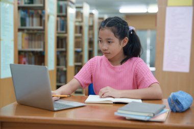 Çevrimiçi eğitim, e-öğrenme. Mutlu Asyalı ilkokul kızı şık günlük kıyafetler içinde, uzaktan çalışıyor, dizüstü bilgisayar kullanıyor, online dersler dinliyor, kütüphanede otururken notlar alıyor..