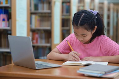 Çevrimiçi eğitim, e-öğrenme. Mutlu Asyalı ilkokul kızı şık günlük kıyafetler içinde, uzaktan çalışıyor, dizüstü bilgisayar kullanıyor, online dersler dinliyor, kütüphanede otururken notlar alıyor..