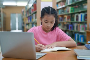 Çevrimiçi eğitim, e-öğrenme. Mutlu Asyalı ilkokul kızı şık günlük kıyafetler içinde, uzaktan çalışıyor, dizüstü bilgisayar kullanıyor, online dersler dinliyor, kütüphanede otururken notlar alıyor..