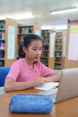 Çevrimiçi eğitim, e-öğrenme. Mutlu Asyalı ilkokul kızı şık günlük kıyafetler içinde, uzaktan çalışıyor, dizüstü bilgisayar kullanıyor, online dersler dinliyor, kütüphanede otururken notlar alıyor..