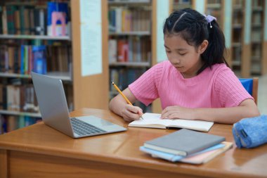 Çevrimiçi eğitim, e-öğrenme. Mutlu Asyalı ilkokul kızı şık günlük kıyafetler içinde, uzaktan çalışıyor, dizüstü bilgisayar kullanıyor, online dersler dinliyor, kütüphanede otururken notlar alıyor..