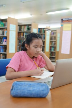 Çevrimiçi eğitim, e-öğrenme. Mutlu Asyalı ilkokul kızı şık günlük kıyafetler içinde, uzaktan çalışıyor, dizüstü bilgisayar kullanıyor, online dersler dinliyor, kütüphanede otururken notlar alıyor..