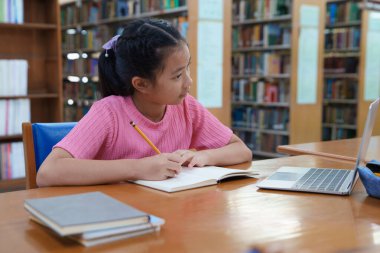 Çevrimiçi eğitim, e-öğrenme. Mutlu Asyalı ilkokul kızı şık günlük kıyafetler içinde, uzaktan çalışıyor, dizüstü bilgisayar kullanıyor, online dersler dinliyor, kütüphanede otururken notlar alıyor..