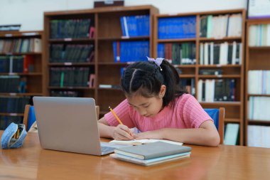 Çevrimiçi eğitim, e-öğrenme. Mutlu Asyalı ilkokul kızı şık günlük kıyafetler içinde, uzaktan çalışıyor, dizüstü bilgisayar kullanıyor, online dersler dinliyor, kütüphanede otururken notlar alıyor..