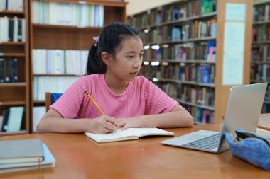 Çevrimiçi eğitim, e-öğrenme. Mutlu Asyalı ilkokul kızı şık günlük kıyafetler içinde, uzaktan çalışıyor, dizüstü bilgisayar kullanıyor, online dersler dinliyor, kütüphanede otururken notlar alıyor..