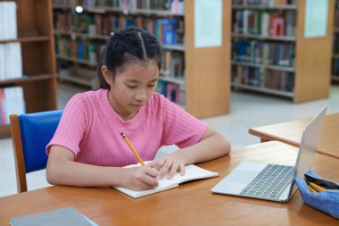 Çevrimiçi eğitim, e-öğrenme. Mutlu Asyalı ilkokul kızı şık günlük kıyafetler içinde, uzaktan çalışıyor, dizüstü bilgisayar kullanıyor, online dersler dinliyor, kütüphanede otururken notlar alıyor..