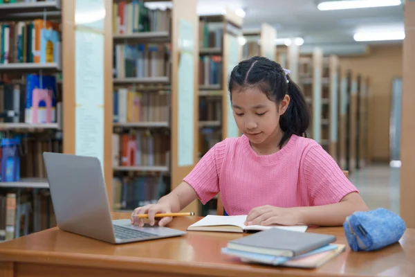 Çevrimiçi eğitim, e-öğrenme. Mutlu Asyalı ilkokul kızı şık günlük kıyafetler içinde, uzaktan çalışıyor, dizüstü bilgisayar kullanıyor, online dersler dinliyor, kütüphanede otururken notlar alıyor..