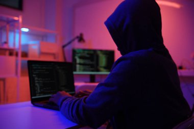 Karanlık bir odada bilgisayar üzerinde çalışan kapüşonlu bir bilgisayar korsanı. Siber güvenlik, hackleme, siber suç ve veri gizliliği tehditleri için bir kavram.