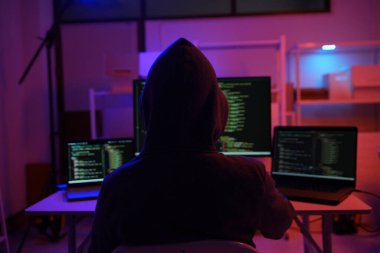 Karanlık bir odada bilgisayar üzerinde çalışan kapüşonlu bir bilgisayar korsanı. Siber güvenlik, hackleme, siber suç ve veri gizliliği tehditleri için bir kavram.