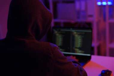 Karanlık bir odada bilgisayar üzerinde çalışan kapüşonlu bir bilgisayar korsanı. Siber güvenlik, hackleme, siber suç ve veri gizliliği tehditleri için bir kavram.