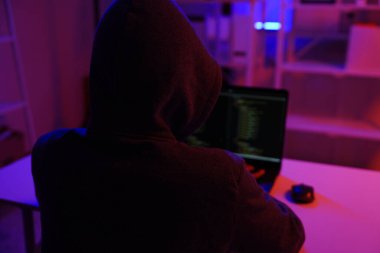 Karanlık bir odada bilgisayar üzerinde çalışan kapüşonlu bir bilgisayar korsanı. Siber güvenlik, hackleme, siber suç ve veri gizliliği tehditleri için bir kavram.
