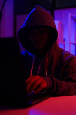 Karanlık bir odada bilgisayar üzerinde çalışan kapüşonlu bir bilgisayar korsanı. Siber güvenlik, hackleme, siber suç ve veri gizliliği tehditleri için bir kavram.