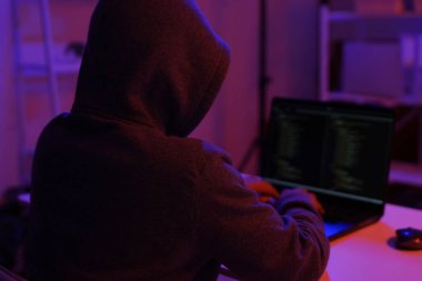 Karanlık bir odada bilgisayar üzerinde çalışan kapüşonlu bir bilgisayar korsanı. Siber güvenlik, hackleme, siber suç ve veri gizliliği tehditleri için bir kavram.