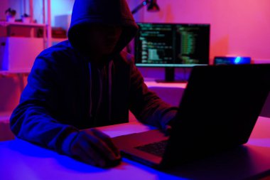 Karanlık bir odada bilgisayar üzerinde çalışan kapüşonlu bir bilgisayar korsanı. Siber güvenlik, hackleme, siber suç ve veri gizliliği tehditleri için bir kavram.