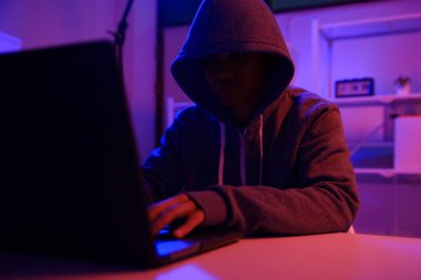 Karanlık bir odada bilgisayar üzerinde çalışan kapüşonlu bir bilgisayar korsanı. Siber güvenlik, hackleme, siber suç ve veri gizliliği tehditleri için bir kavram.
