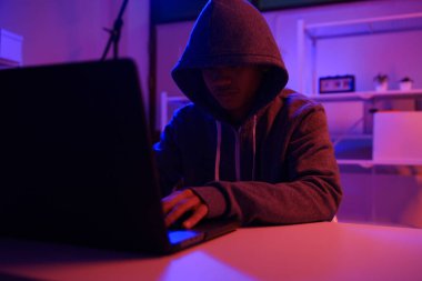 Karanlık bir odada bilgisayar üzerinde çalışan kapüşonlu bir bilgisayar korsanı. Siber güvenlik, hackleme, siber suç ve veri gizliliği tehditleri için bir kavram.