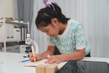 Sınıfta mikroskoptan gözlemleri kaydeden genç bir kız öğrenci. BTMM eğitimi, erken veri analizi ve çocuklar için araştırma becerileri kavramı.