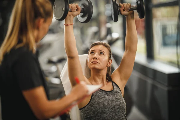 体育館で女性のパーソナルトレーナーと仕事をするスポーツウェアで筋肉質の若い女性のショット 彼女はダンベルと彼女の筋肉を汲み上げている — ストック写真