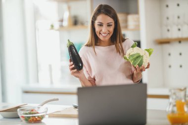 Genç bir kadının evde sağlıklı bir yemek hazırlarken bilgisayarı kullanarak video bloğu hazırlaması..