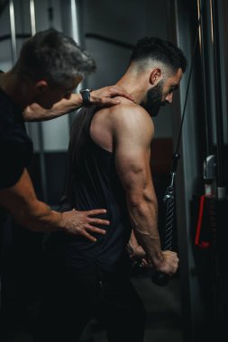 Spor giyim bölümünde spor salonunda kişisel antrenörle çalışan kaslı bir adamı vurmuşlar. Güç antrenmanı sırasında kablo trisepsleri çekiyor..