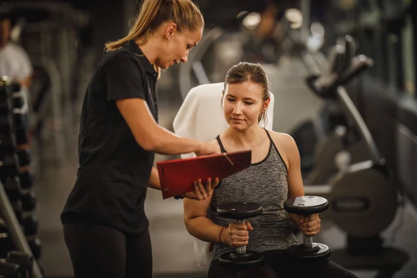 体育館で女性のパーソナルトレーナーと仕事をするスポーツウェアで筋肉質の若い女性のショット 彼女はダンベルと彼女の筋肉を汲み上げている — ストック写真
