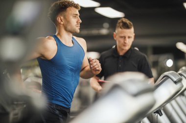 Spor salonundaki koşu bandında koşan kaslı bir gencin fotoğrafı. Kişisel antrenörle çalışıyor..