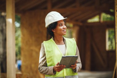 Afrikalı bir kadın mimarın dijital tablet kullanarak ve yeni ahşap bir evin şantiyesini kontrol ederken görüntüsü. Koruyucu iş kıyafeti ve beyaz kask giyiyor..
