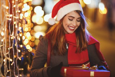 Kırmızı hediye kutusu olan gülümseyen genç bir kadının Noel zamanı şehir caddesinde eğlendiğini gösteren bir fotoğraf..