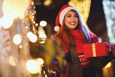 Kırmızı hediye kutusu olan neşeli genç bir kadının çekimi Noel zamanı şehir sokaklarında eğleniyor..