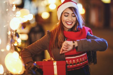 Kırmızı hediye kutusu olan mutlu genç bir kadının çekimleri var. Saatine bakıyor ve Noel zamanı şehir caddesinde birini bekliyor..