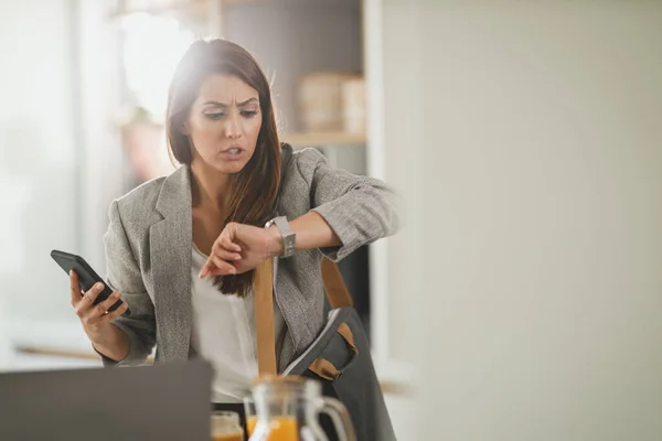 Shot of a multi-tasking young business woman looking on watch and using smartphone in her kitchen while getting ready to go to work.