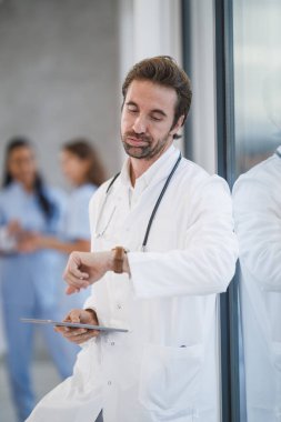 Covid-19 salgını sırasında, bir pencerenin yanında durup hastane koridorundaki saate bakarken bitkin görünen bir doktorun görüntüsü..