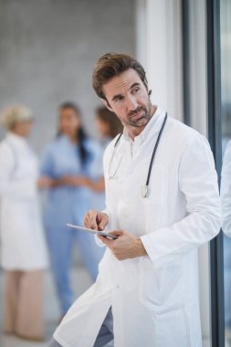 Olgun bir doktorun dijital tablet kullanırken ve hastane koridorunda hızlı bir mola verirken pencereden dışarı bakarken görüntüsü..