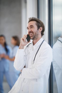 Bir hastane koridorunda durup, akıllı bir telefonla konuşan gülümseyen bir doktor..