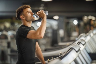 Spor salonunda koşu bandında koşan bir gencin fotoğrafı. Su içebiliyor..