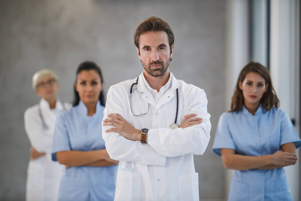 Portrait of a confident doctor standing with arms crossed in a hospital hallway with his colleagues in the background.