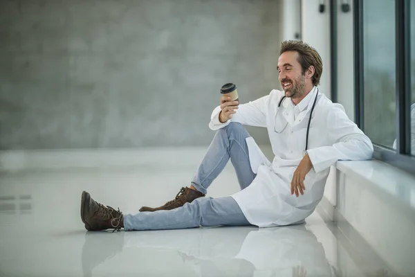 Médico Sonriente Sentado Suelo Cerca Una Ventana Mientras Toma Café — Foto de Stock