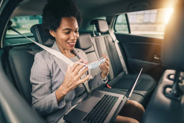 Shot of a successful black businesswoman with protective mask using laptop while sitting in the backseat of a car on her morning commute during COVID-19 pandemic.