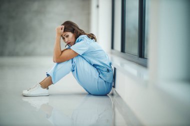 Stresli genç bir hemşire, Covid-19 salgını sırasında boş bir hastane koridorunda kısa bir mola verirken pencerenin yanında oturuyor..