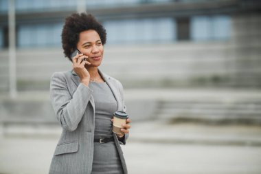 Başarılı bir siyahi iş kadını, bir şirket binasının önünde kahve molası verirken akıllı bir telefonda uygulama kullanıyor..