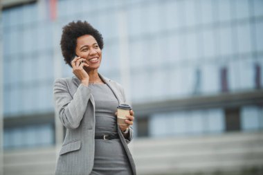 Başarılı bir siyahi iş kadını, bir şirket binasının önünde kahve molası verirken akıllı bir telefonda uygulama kullanıyor..