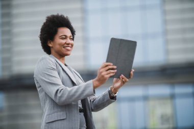 Şirket binasının önünde kısa bir aradan sonra dijital tabletle video görüşmesi yapan gülümseyen zenci bir iş kadını portresi..