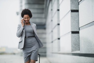 Bir şirket binasının önünde yürürken akıllı telefon kullanan gülümseyen zenci bir iş kadını..
