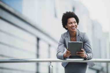 Şirket binasının önünde dijital tablet kullanan gülümseyen zenci bir iş kadını..