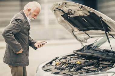 Endişeli kıdemli iş adamı akıllı telefon kullanıyor ve yol gezisinde arabasının kaputunun altına bakıyor..